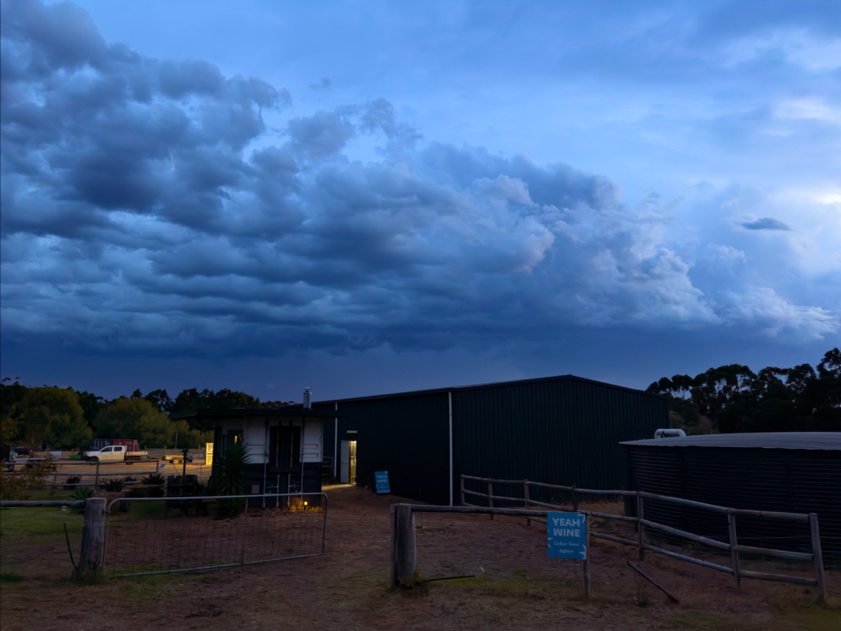 Dark, stormy clouds hang over a low farm building at dusk, with a gate and fenced dirt entry in the foreground. A blue sign near the fence reads “YEAH WINE,” and a large black water tank sits to the right.
