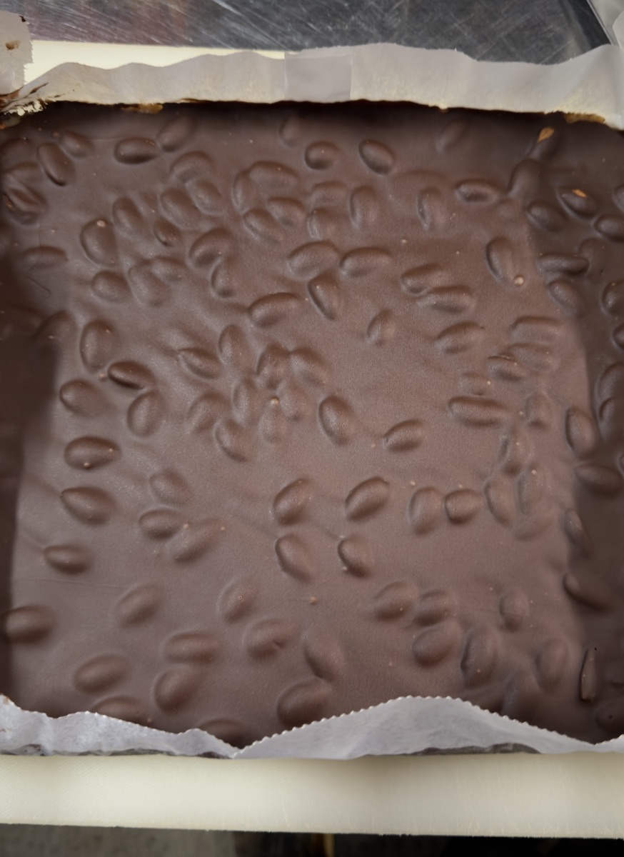 Close-up of a rectangular slab of dark chocolate sprinkled with small oval nuts or seeds. The chocolate sits on white parchment paper with torn edges.
