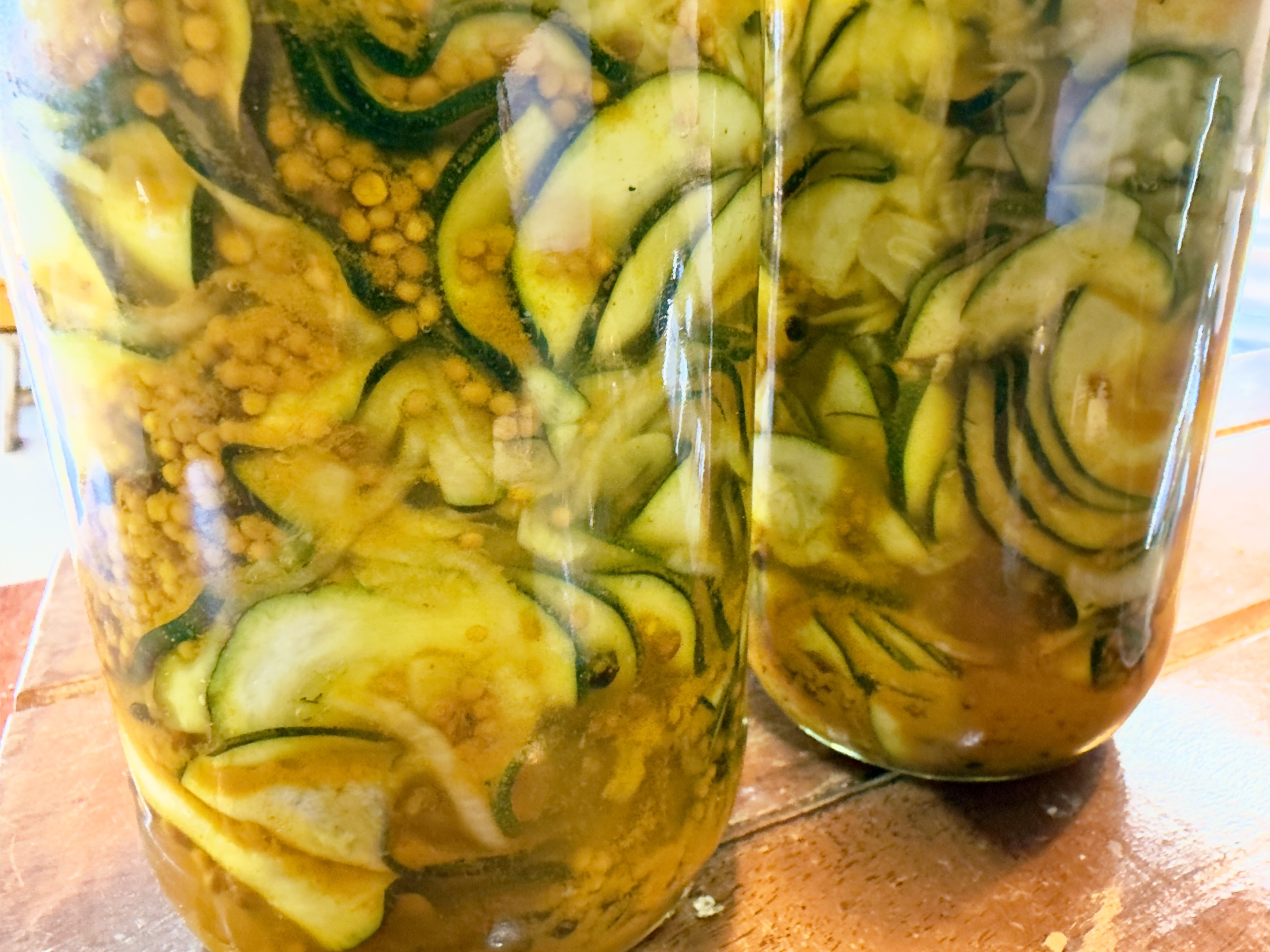 Two glass jars filled with thinly sliced cucumbers in yellow-green brine, with mustard seeds visible. The jars sit on a wooden surface, with one jar in the foreground and another slightly behind it.
