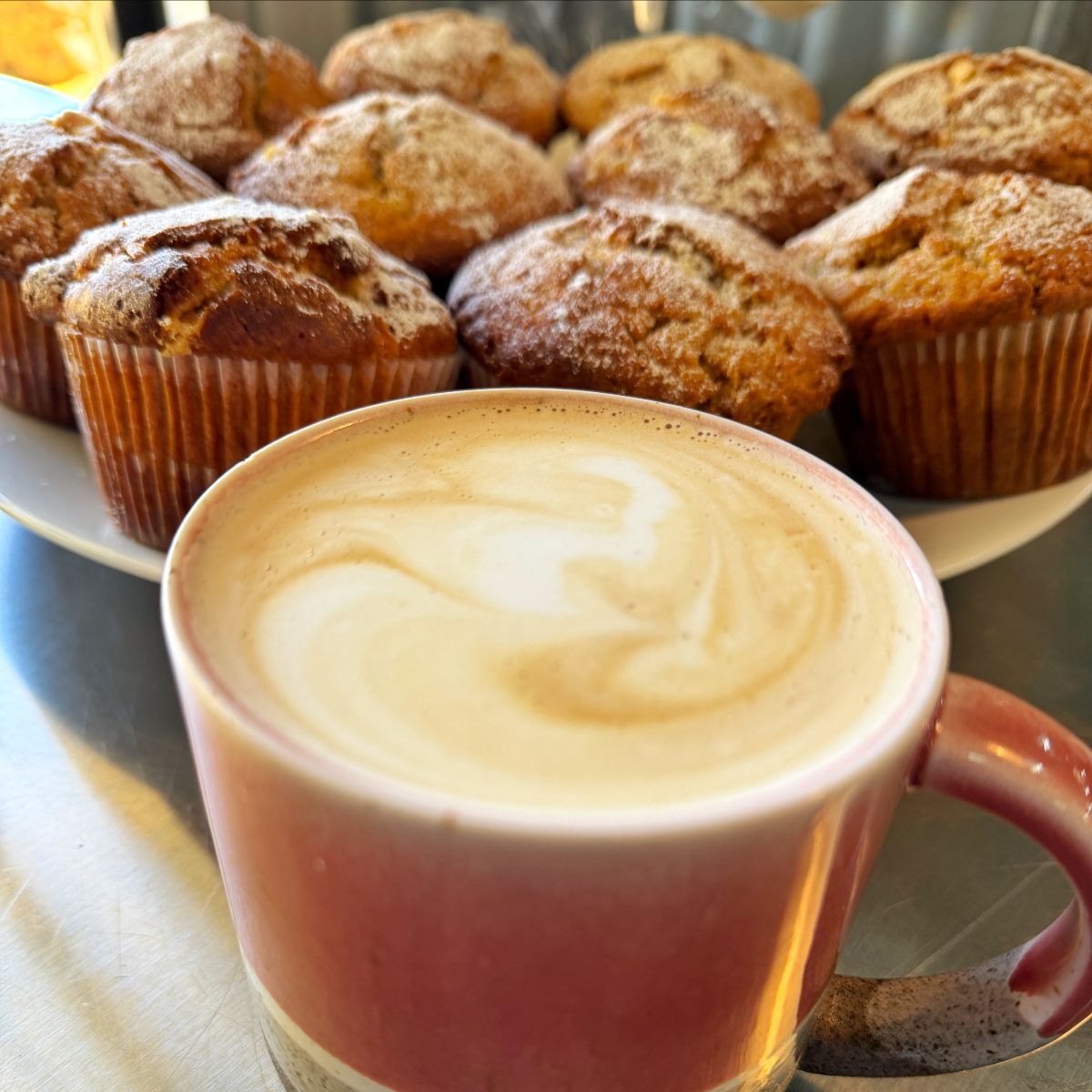 Pink mug of cappuccino with frothy latte art in the foreground. Behind it, a plate piled with sugar-dusted muffins on a metal counter.