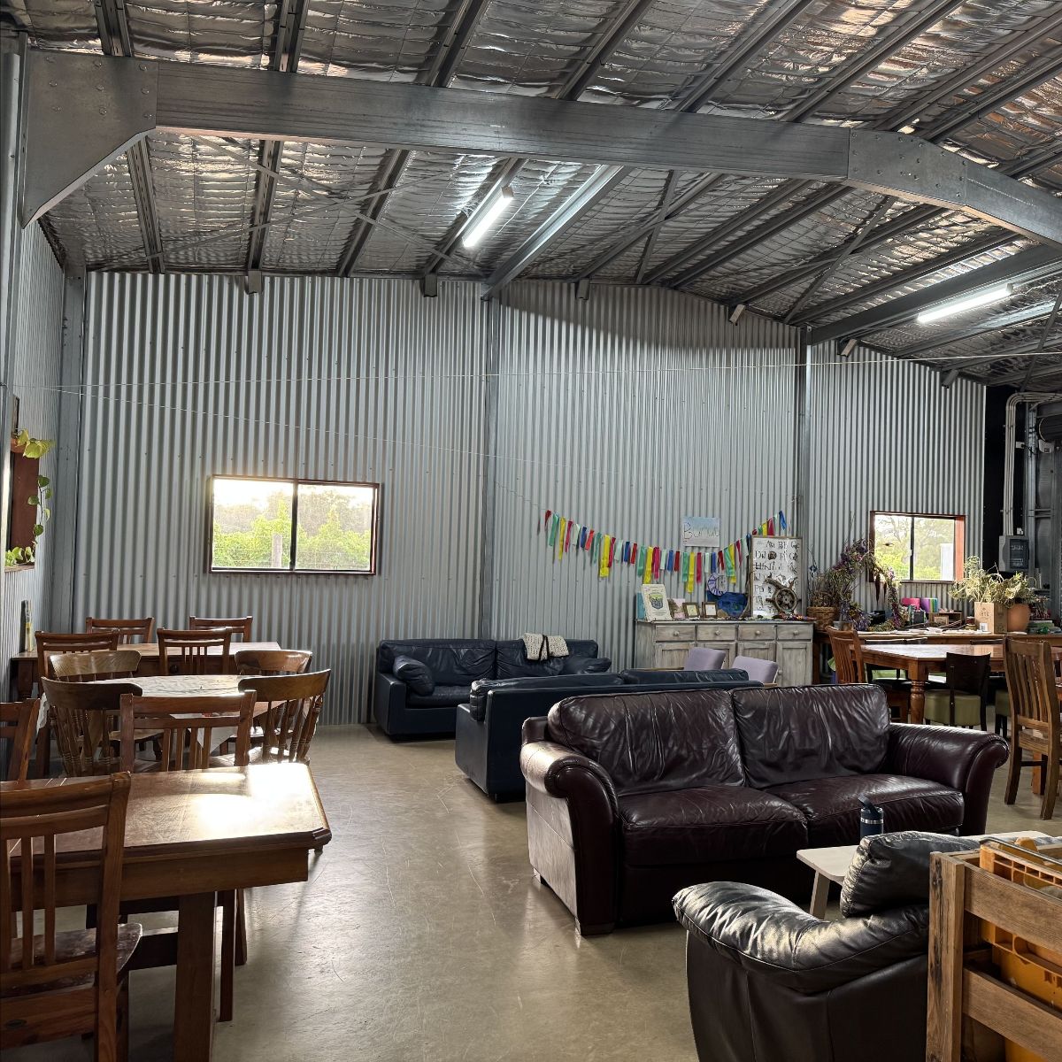 Open shed-like hall with corrugated metal walls and a high insulated ceiling, furnished with wooden tables and chairs and several leather couches. A string of colorful bunting hangs above a sideboard, with plants and two windows along the walls.