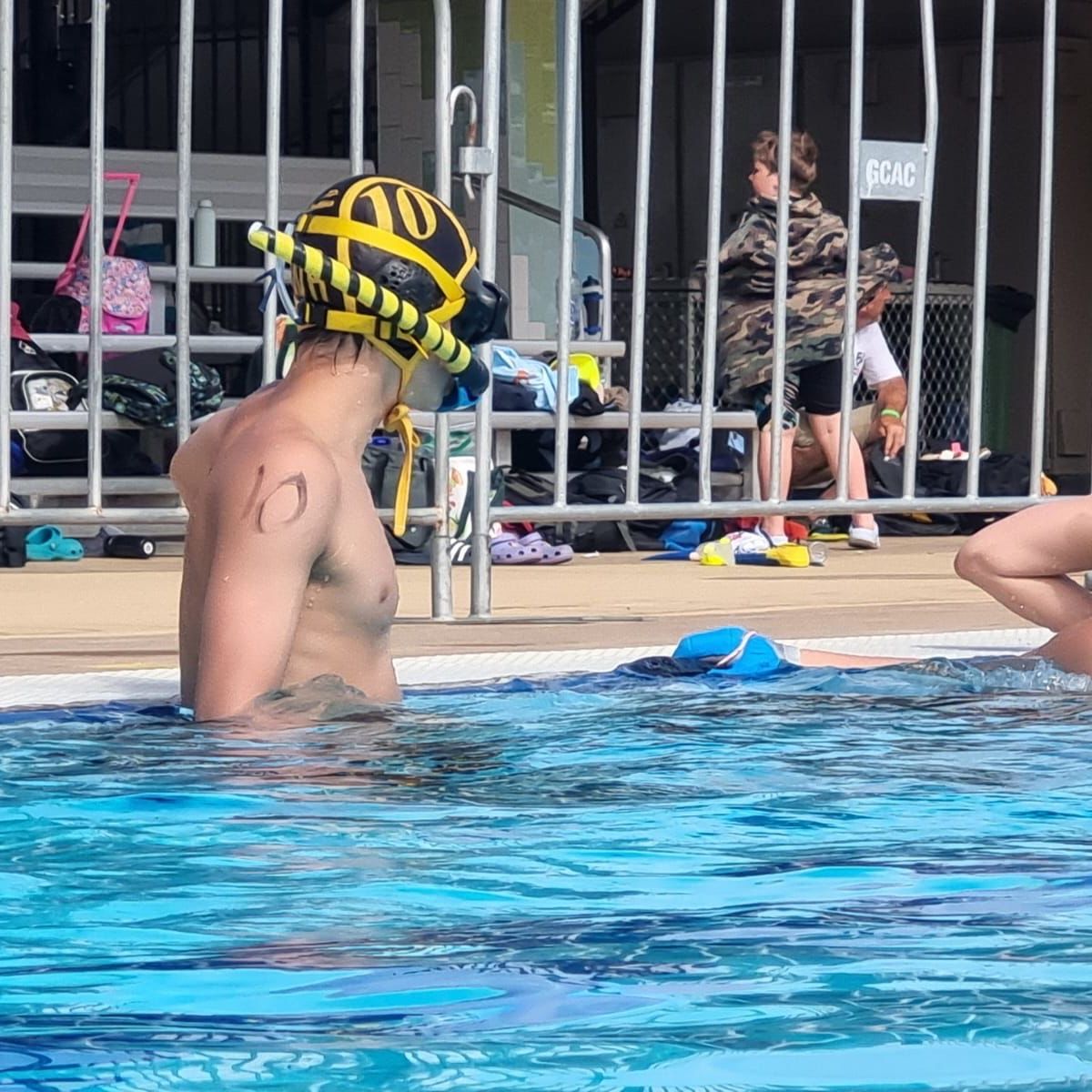 Water polo player in a pool wearing a black-and-yellow cap labeled “20” and a snorkel, with one arm resting on the surface. Pool deck with metal railings and scattered bags and shoes in the background.