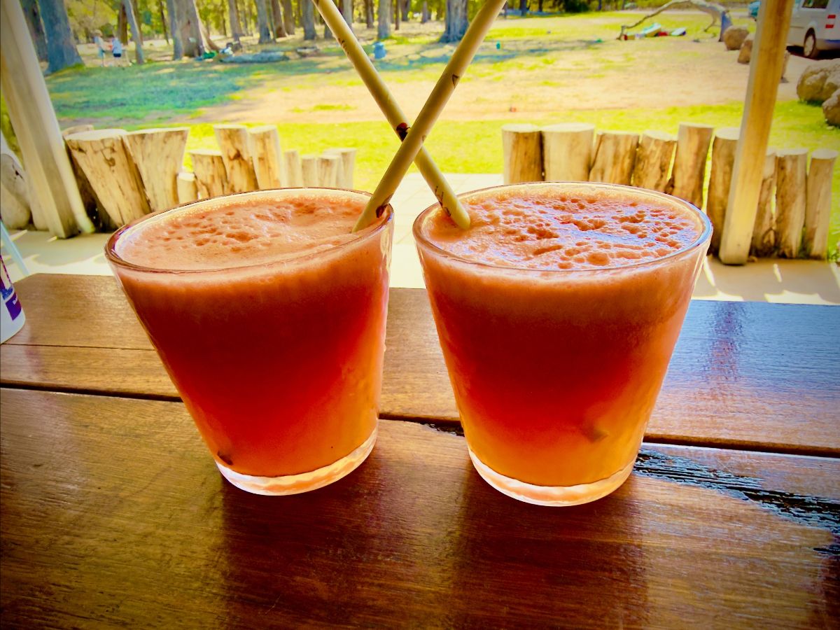 Two short glasses of pink-orange smoothie sit on a wooden table, each with a straw angled inward so the straws cross. In the background are sunlit grass, trees, and a rustic wooden fence.