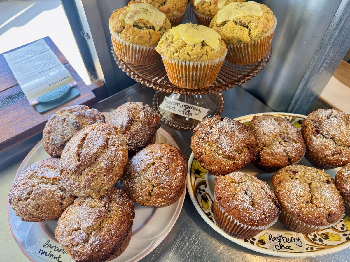 Assorted muffins displayed on plates and a raised glass stand inside a metal cabinet, including banana walnut and raspberry choc varieties. A handwritten label under the stand reads “mushroom, rosemary, rocket + feta.”