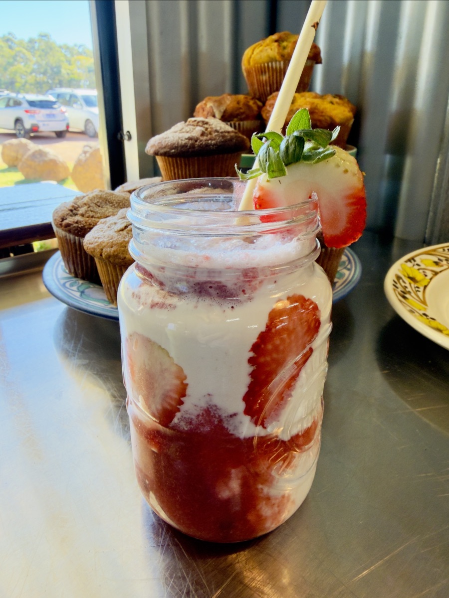 Mason jar filled with a pink-and-white strawberry drink, with sliced strawberries visible inside and a whole strawberry on the rim beside a straw. Muffins on a plate sit behind it on a metal counter near a window with parked cars outside.