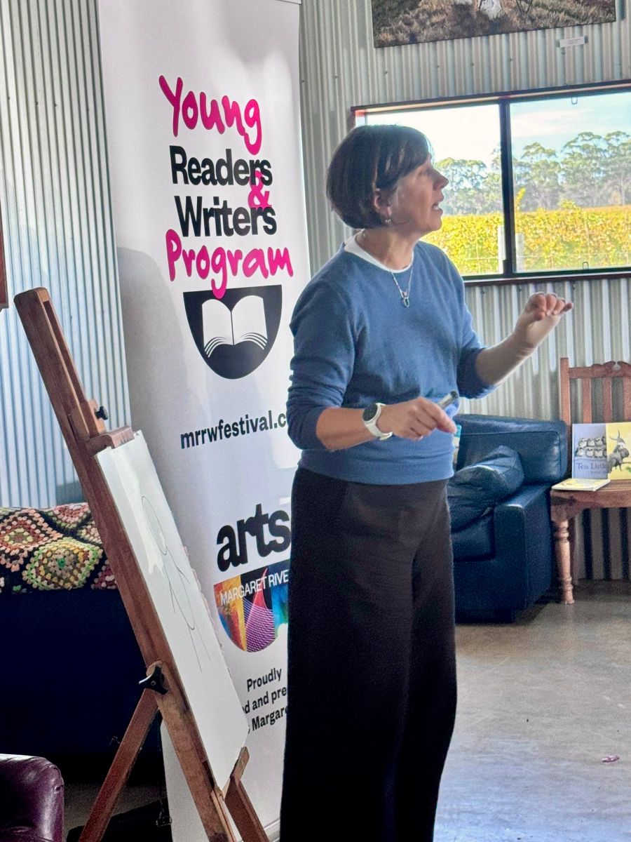 Woman stands indoors gesturing beside an easel with a large sheet of paper, as if speaking to a group. A banner behind her reads “Young Readers & Writers Program,” and a window shows green fields outside.