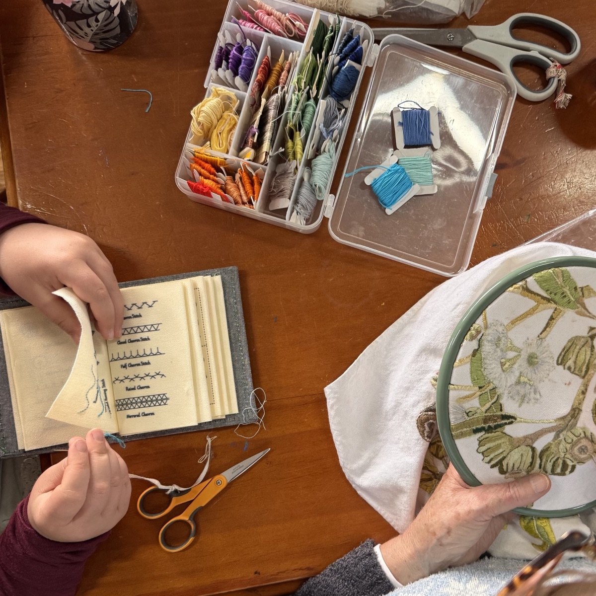 Hands at a wooden table with embroidery supplies, including a plastic organizer of colorful thread, scissors, and a stitch-sample booklet. An embroidery hoop holds fabric with a partly stitched floral design.