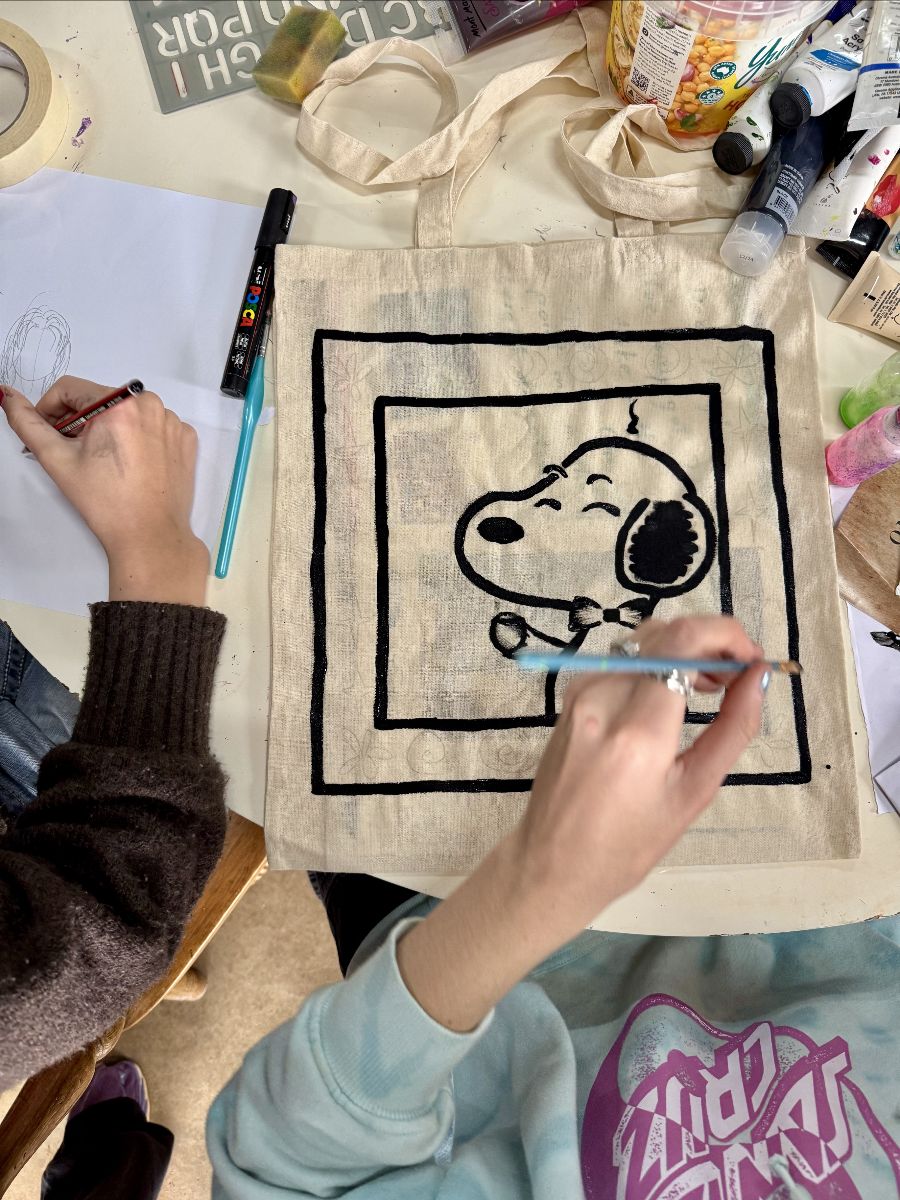 Hands at a craft table draw on paper and paint a black-outlined Snoopy design on a plain canvas tote bag. Paint tubes, markers, and brushes are scattered around the bag.