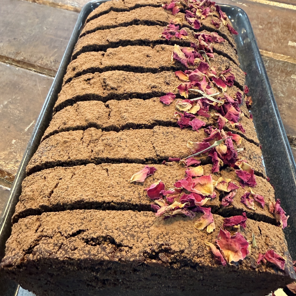 Sliced chocolate loaf cake on a black tray, dusted with cocoa powder. Dried rose petals are scattered along the top.