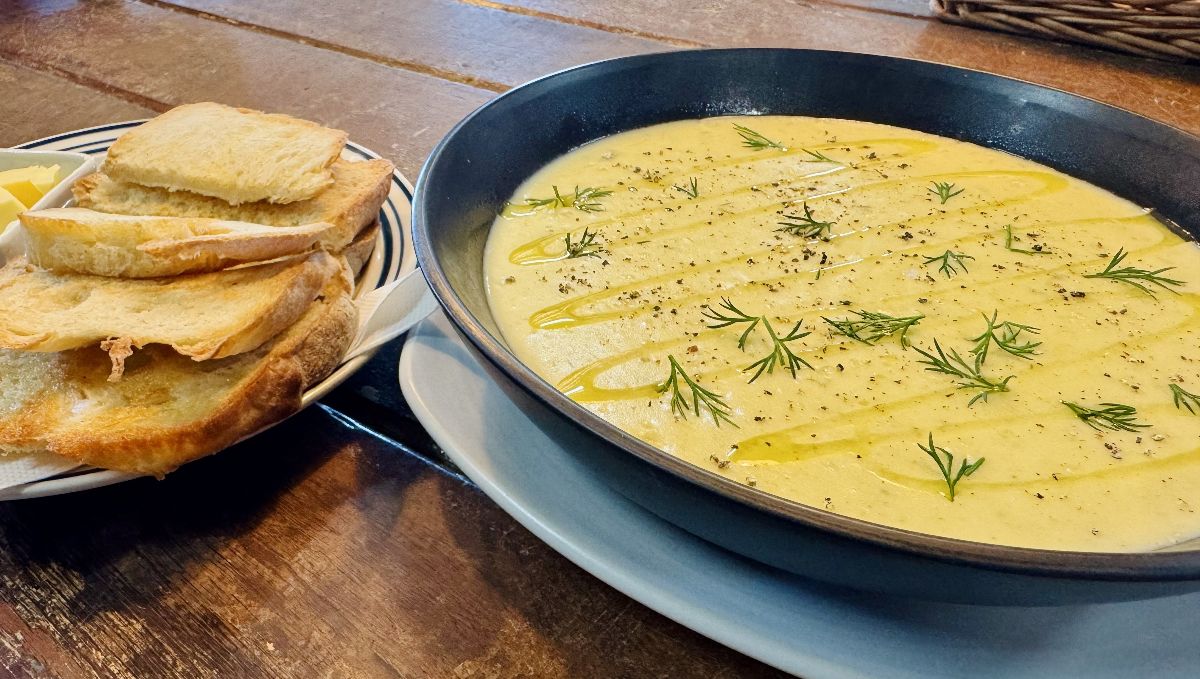 Large shallow bowl of creamy yellow soup drizzled with oil and topped with black pepper and dill sprigs on a wooden table. Plate of sliced bread and a small dish of butter sit beside it.