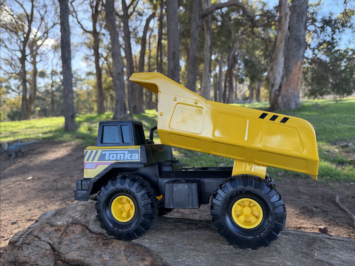 Yellow and black Tonka toy dump truck with its bed raised, sitting on a log. Blurred background shows a wooded park with tall trees and a dirt path.