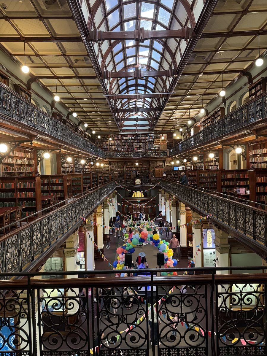 Two-level library hall with ornate iron railings, tall bookshelves, and a glass-vaulted ceiling viewed from an upper balcony. Colorful bunting and a balloon arch decorate the ground floor where several people are walking around.