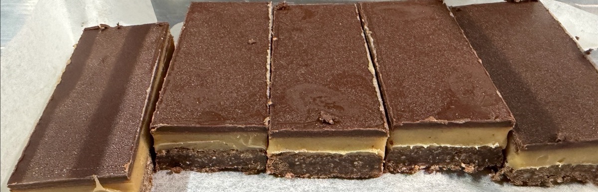 Several rectangular dessert bars with a dark chocolate top and pale middle layer sit on a metal tray, some cut into portions. Crumbly dark base is visible along the bottom edges.