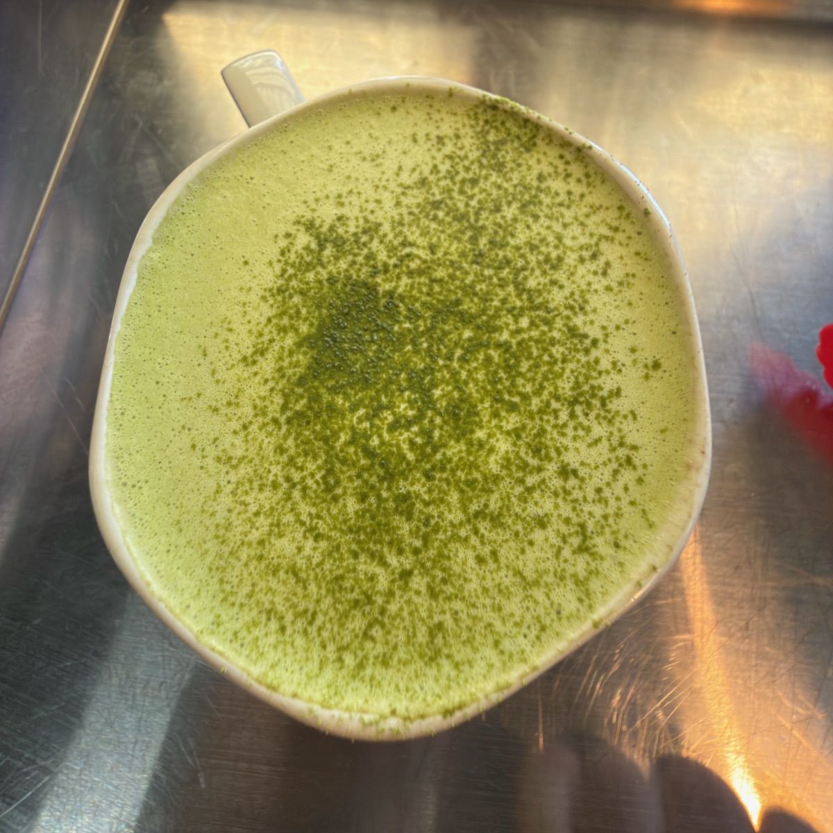 Overhead view of a white mug filled with pale green foam, topped with darker green powder speckled across the surface. The mug sits on a stainless-steel counter with warm reflections.