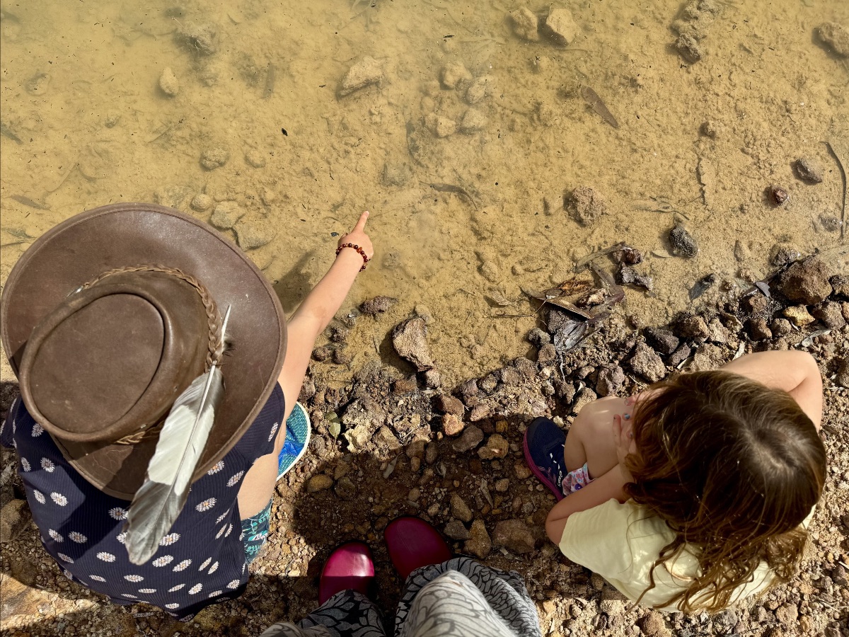 Two children sit on a rocky shore beside shallow, clear water; one wears a brown hat with a feather and points into the water. An adult’s legs and pink shoes are visible at the bottom edge.