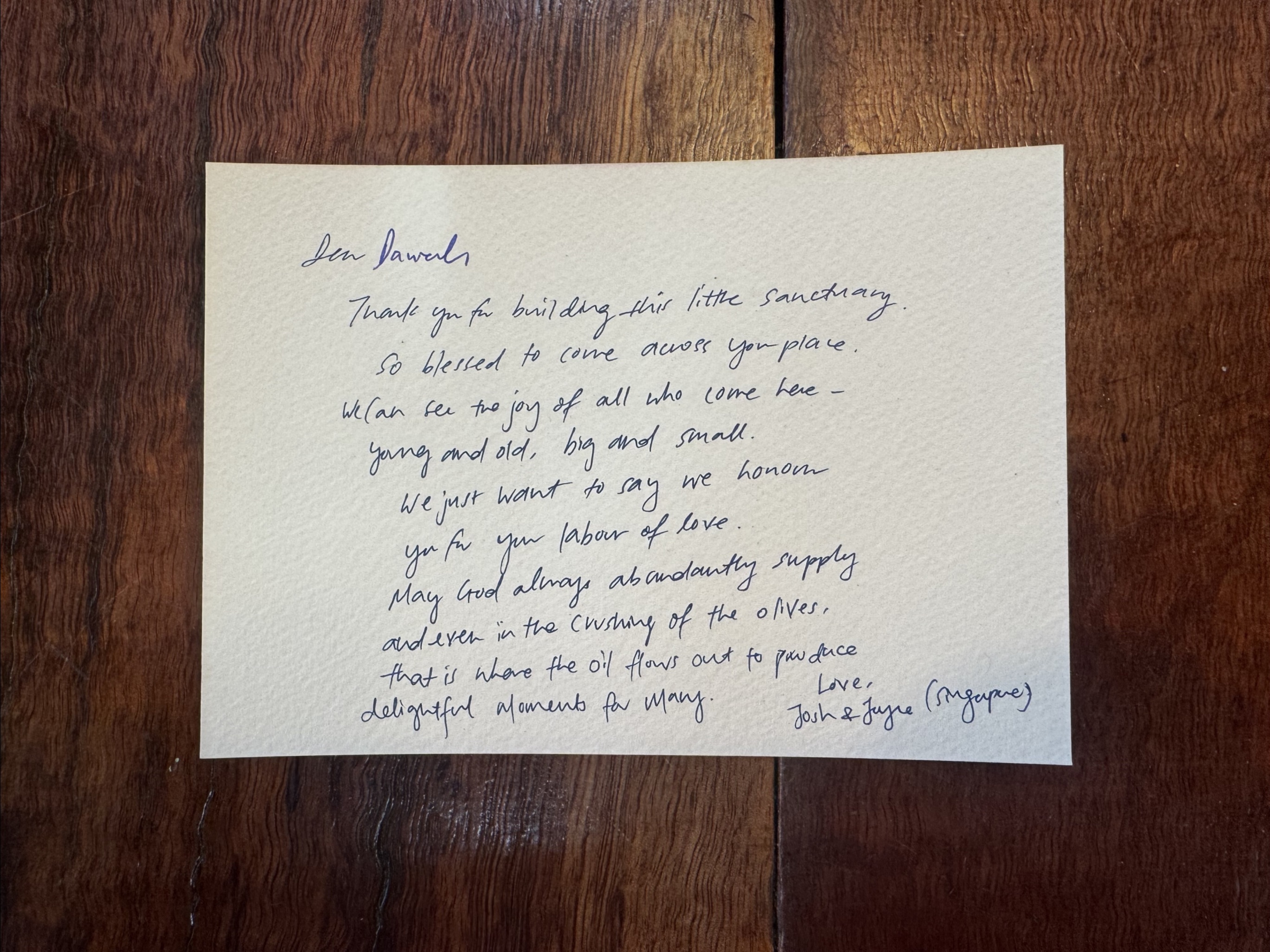 Handwritten thank-you note in blue ink on cream paper, laid on a dark wooden tabletop. The note begins “Dear Pamela” and is signed “Josh & Joyce (Singapore)” in the bottom right.