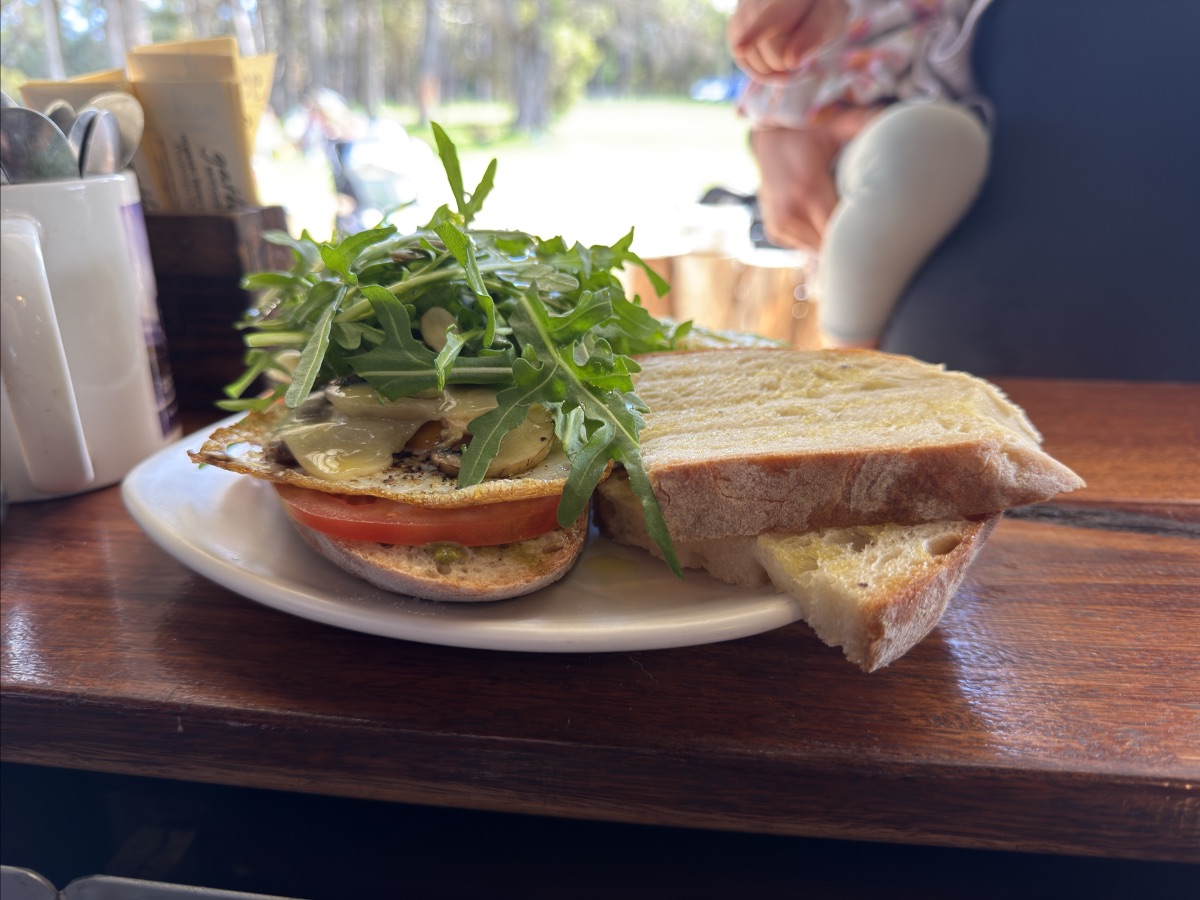 Open sandwich on a white plate with tomato slices, melted cheese, and a handful of arugula, served with a thick slice of bread on a wooden table. Condiment containers and sugar packets sit beside it, with a softly blurred outdoor background.